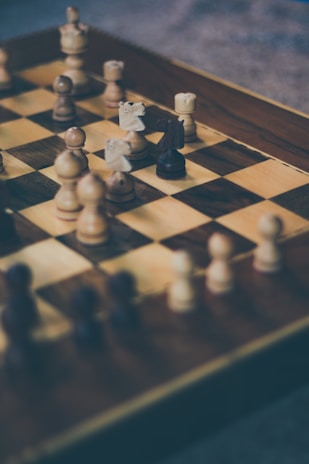 A close-up view of a wooden chessboard with several chess pieces arranged in mid-game. The focus is on the pieces in the middle of the board, with a mix of light and dark pieces visible. The background is slightly blurred, emphasizing the chessboard.