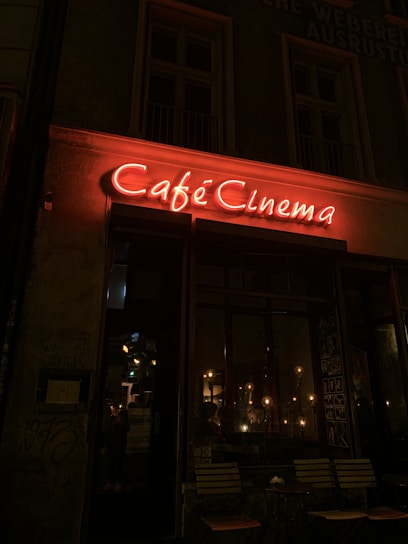 A neon sign in red illuminates the words 'Café Cinema' above the entrance of a dimly lit cafe. The warm glow of interior lighting is visible through the large windows, creating a cozy and inviting atmosphere. There are three sets of wooden chairs and tables outside the cafe.