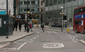 A person is riding a bicycle on a city street with marked bike lanes. Several pedestrians are visible on the sidewalk to the left, while a red double-decker bus can be seen on the right side of the street. The area is surrounded by modern glass buildings, and street signs and traffic lights are present.