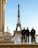 Three police officers in riot gear stand in front of the Eiffel Tower on a clear day. Behind them, several statues and birds are visible. The sky is blue, and the scene takes place on a paved area with modern and classical architecture elements.
