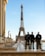 Three police officers in riot gear stand in front of the Eiffel Tower on a clear day. Behind them, several statues and birds are visible. The sky is blue, and the scene takes place on a paved area with modern and classical architecture elements.