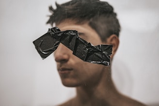 A person stands in front of a blurred background with black tape obscuring their eyes. The tape is crumpled and unevenly applied, creating a textured appearance. The person's features are partially visible, with focus on the obscured eyes.