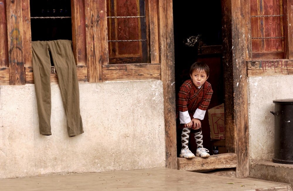 boy in red and black plaid dress shirt and brown pants sitting on brown wooden door