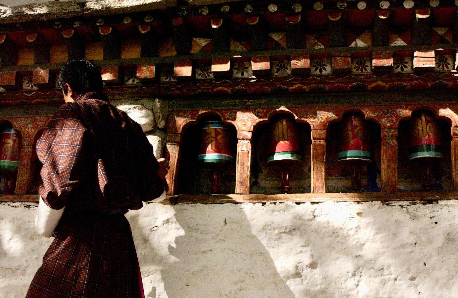 A worshipper in traditional checked dress turning a row of red prayer wheels set into a whitewashed temple wall