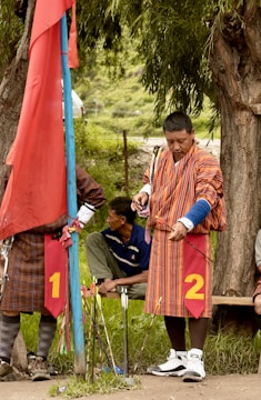 A man dressed in traditional striped attire is preparing to shoot an arrow, standing near a blue pole with a red flag. Another man is sitting behind him on a bench. They are in an outdoor setting with greenery and trees around.