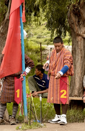 A man dressed in traditional striped attire is preparing to shoot an arrow, standing near a blue pole with a red flag. Another man is sitting behind him on a bench. They are in an outdoor setting with greenery and trees around.