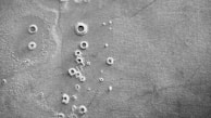 A black and white aerial view of a cratered, rocky surface resembling that of the moon or another celestial body. The texture appears rough with various sizes of circular indentations scattered throughout.