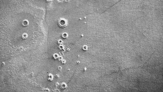 A black and white aerial view of a cratered, rocky surface resembling that of the moon or another celestial body. The texture appears rough with various sizes of circular indentations scattered throughout.