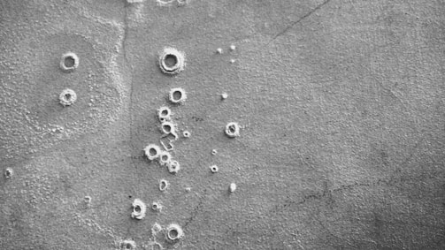 A black and white aerial view of a cratered, rocky surface resembling that of the moon or another celestial body. The texture appears rough with various sizes of circular indentations scattered throughout.