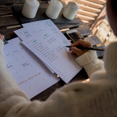 A person in a cozy sweater is writing on a to-do list or planner placed on a wooden table. The setting includes candles, a smartphone, and papers, suggesting an organized and serene atmosphere.