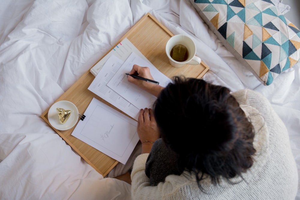 person writing on white paper beside white ceramic mug on white textile