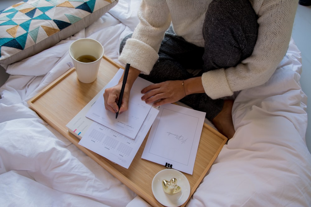 person in white sweater writing on white paper beside white ceramic mug on brown wooden table