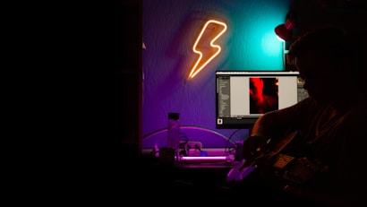 A dimly lit room with neon lighting features a person playing a guitar near a computer desk. A glowing lightning bolt wall light adds to the mood, while the computer screen displays an image with a red and black contrast. Various cables and a water bottle are visible on the desk, contributing to a creative studio-like atmosphere.