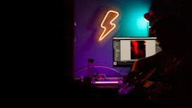 A dimly lit room with neon lighting features a person playing a guitar near a computer desk. A glowing lightning bolt wall light adds to the mood, while the computer screen displays an image with a red and black contrast. Various cables and a water bottle are visible on the desk, contributing to a creative studio-like atmosphere.