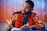 A man wearing a brightly colored patterned jacket is sitting at a table. He is holding a pen over a notebook, appearing thoughtful or focused. The background consists of vertical blinds, creating a warm ambiance.