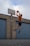 A person dressed in athletic wear is jumping towards a basketball hoop mounted on a brick wall. They are holding a basketball with one hand and appear to be about to make a shot. The background consists of a brick wall with two blue vertical garage doors and a clear sky with some clouds.