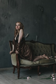 A woman with long hair, wearing a lace outfit and a silk robe, poses on a vintage, green sofa in a dimly lit room. The walls are textured and dark, with a slightly distressed look. A few pink flowers are scattered on the floor, adding a touch of color to the otherwise muted scene.