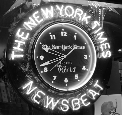 A neon clock with the words 'THE NEW YORK TIMES' and 'NEWSBEAT' illuminated around it. The clock face features the phrase 'expect the World' along with numerals in a classic font. The image is in black and white.