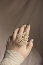 A delicate hand with green-painted nails gently holds small, white baby's breath flowers. The background is softly blurred, creating an intimate and serene atmosphere. The person is wearing a knitted, beige sweater, adding a cozy texture to the overall composition.