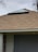 A building with a triangular skylight positioned at the center of a large, shingled roof. The roof is a light brown color and the building has a stone column with a closed gray shutter door below the roofline. A portion of a tree with bare branches is visible against the gray sky in the background.