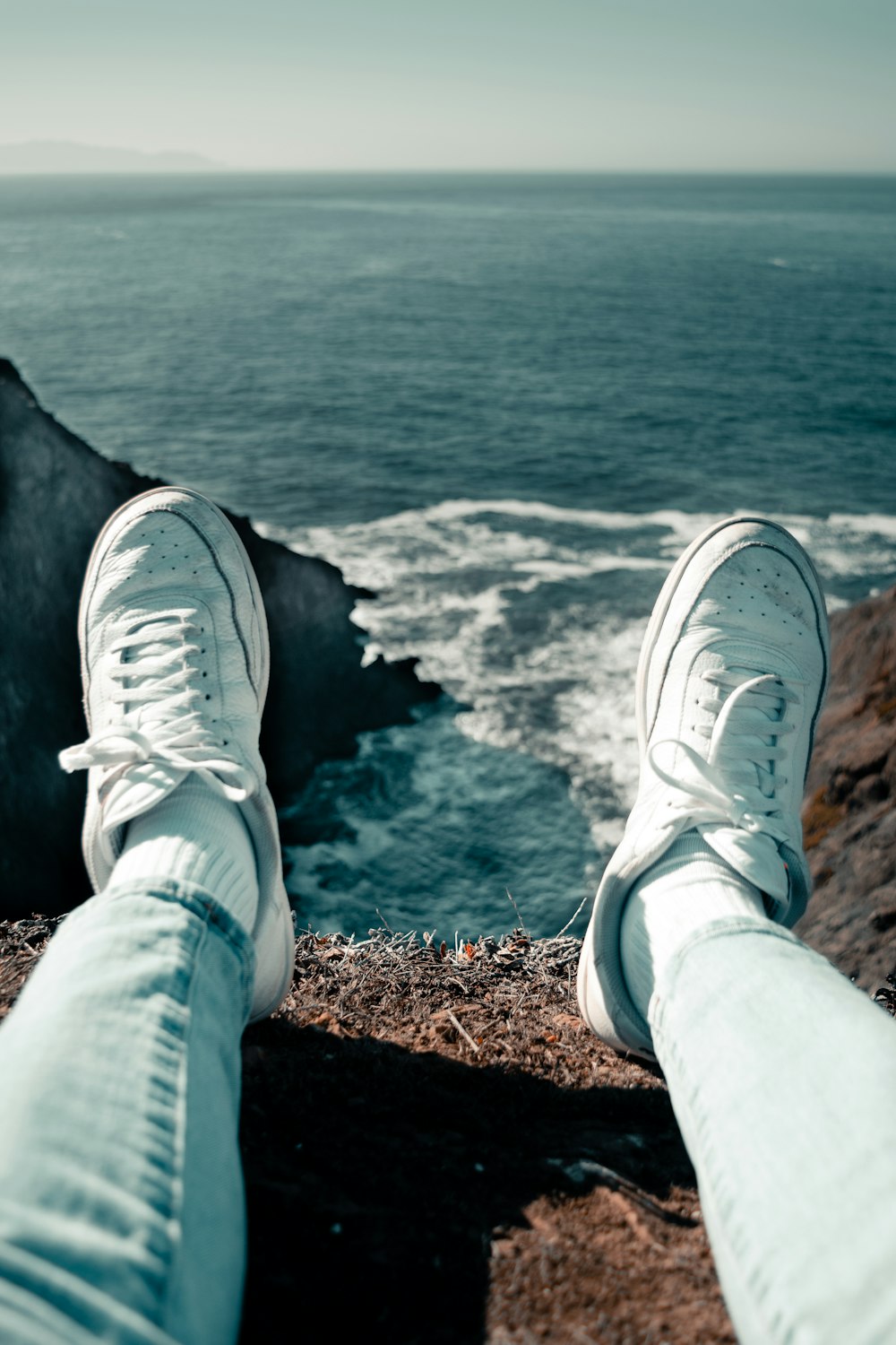 person in white nike sneakers sitting on brown rock near body of water during daytime