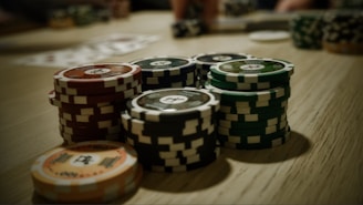 Stacks of poker chips are arranged on a wooden table, with various denominations and colors such as red, green, and black. Some playing cards are blurred in the background, indicating a game setting.
