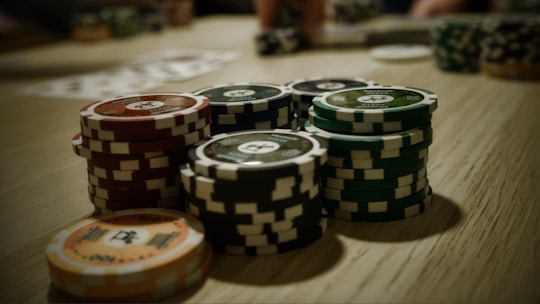 Stacks of poker chips are arranged on a wooden table, with various denominations and colors such as red, green, and black. Some playing cards are blurred in the background, indicating a game setting.