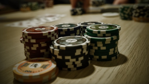 Stacks of poker chips are arranged on a wooden table, with various denominations and colors such as red, green, and black. Some playing cards are blurred in the background, indicating a game setting.