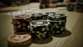 Stacks of poker chips are arranged on a wooden table, with various denominations and colors such as red, green, and black. Some playing cards are blurred in the background, indicating a game setting.