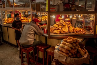 A food stall in a market displays a variety of meats under warm lighting. A man is seated on a stool eating, while a younger person wearing a mask stands nearby. A pile of bread is prominently visible in the foreground. The setting appears dimly lit, with a cozy and bustling atmosphere.