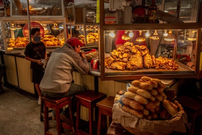 A food stall in a market displays a variety of meats under warm lighting. A man is seated on a stool eating, while a younger person wearing a mask stands nearby. A pile of bread is prominently visible in the foreground. The setting appears dimly lit, with a cozy and bustling atmosphere.