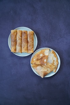 Two plates are set against a dark textured background. One plate contains four neatly arranged spring rolls, and the other has three folded crepes with a golden-brown surface, each showing subtle hints of crispness.