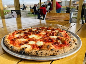 A freshly baked pepperoni pizza sits on a wooden table, its crust perfectly browned and topped with melted cheese and slices of pepperoni. In the background, people are seated at an outdoor dining area under a wooden shelter, with a blurred view of a garden setting.