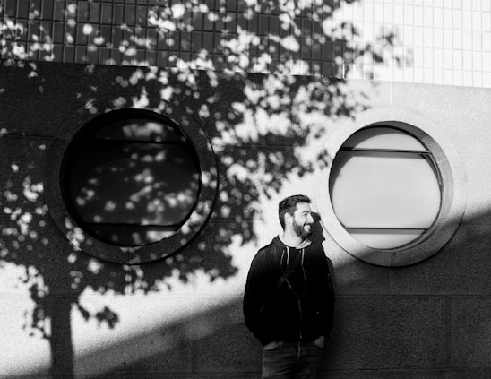 A casually dressed man leaning against a sunlit urban wall, wearing neutral-toned trendy clothes with a relaxed yet confident expression.