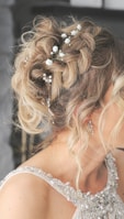 A close-up view of a woman's intricately styled hair adorned with small white flowers and decorative pearls. The hair is blonde and arranged in an elegant, tousled updo. The woman is wearing a sleeveless dress with detailed embellishments, including pearls and crystals. Her earrings are delicate and shiny, complementing the elegant look.