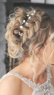 A close-up view of a woman's intricately styled hair adorned with small white flowers and decorative pearls. The hair is blonde and arranged in an elegant, tousled updo. The woman is wearing a sleeveless dress with detailed embellishments, including pearls and crystals. Her earrings are delicate and shiny, complementing the elegant look.