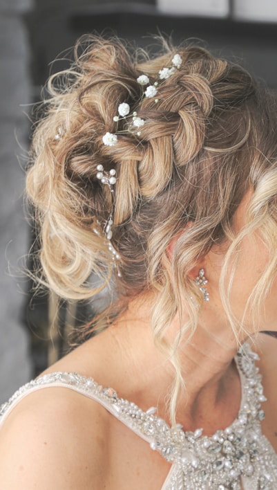 A close-up view of a woman's intricately styled hair adorned with small white flowers and decorative pearls. The hair is blonde and arranged in an elegant, tousled updo. The woman is wearing a sleeveless dress with detailed embellishments, including pearls and crystals. Her earrings are delicate and shiny, complementing the elegant look.