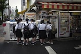 A group of five schoolgirls, wearing uniforms with black skirts and white shirts, are gathered around a row of vending machines in a bustling urban area. They appear to be interacting with the machines, possibly purchasing items. The area is vibrant, with colorful signs and posters adorning the machines and the store front. Pedestrians and city buildings are visible in the background.