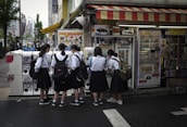 A group of five schoolgirls, wearing uniforms with black skirts and white shirts, are gathered around a row of vending machines in a bustling urban area. They appear to be interacting with the machines, possibly purchasing items. The area is vibrant, with colorful signs and posters adorning the machines and the store front. Pedestrians and city buildings are visible in the background.