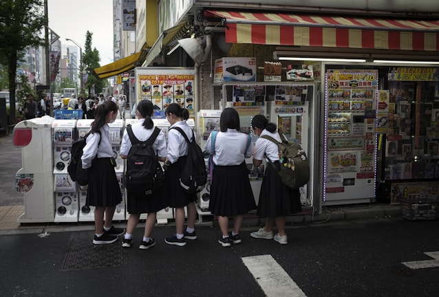A group of five schoolgirls, wearing uniforms with black skirts and white shirts, are gathered around a row of vending machines in a bustling urban area. They appear to be interacting with the machines, possibly purchasing items. The area is vibrant, with colorful signs and posters adorning the machines and the store front. Pedestrians and city buildings are visible in the background.