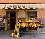 A small, charming grocery store with an awning labeled 'ALIMENTARI Franca' is showcased. Various fresh fruits such as bananas, oranges, apples, and pears are neatly arranged in black crates outside the store. A bright yellow hand truck with red handles is placed in front. The shop entrance displays wooden doors with glass panes and is decorated with boards listing local products.