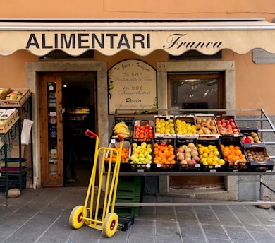 A small, charming grocery store with an awning labeled 'ALIMENTARI Franca' is showcased. Various fresh fruits such as bananas, oranges, apples, and pears are neatly arranged in black crates outside the store. A bright yellow hand truck with red handles is placed in front. The shop entrance displays wooden doors with glass panes and is decorated with boards listing local products.