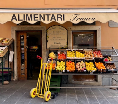 A small, charming grocery store with an awning labeled 'ALIMENTARI Franca' is showcased. Various fresh fruits such as bananas, oranges, apples, and pears are neatly arranged in black crates outside the store. A bright yellow hand truck with red handles is placed in front. The shop entrance displays wooden doors with glass panes and is decorated with boards listing local products.