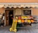 A small, charming grocery store with an awning labeled 'ALIMENTARI Franca' is showcased. Various fresh fruits such as bananas, oranges, apples, and pears are neatly arranged in black crates outside the store. A bright yellow hand truck with red handles is placed in front. The shop entrance displays wooden doors with glass panes and is decorated with boards listing local products.