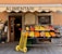 A small, charming grocery store with an awning labeled 'ALIMENTARI Franca' is showcased. Various fresh fruits such as bananas, oranges, apples, and pears are neatly arranged in black crates outside the store. A bright yellow hand truck with red handles is placed in front. The shop entrance displays wooden doors with glass panes and is decorated with boards listing local products.