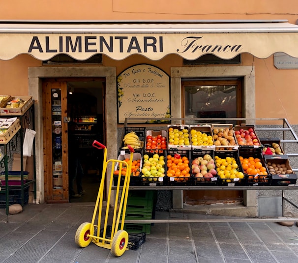 A small, charming grocery store with an awning labeled 'ALIMENTARI Franca' is showcased. Various fresh fruits such as bananas, oranges, apples, and pears are neatly arranged in black crates outside the store. A bright yellow hand truck with red handles is placed in front. The shop entrance displays wooden doors with glass panes and is decorated with boards listing local products.