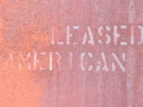 The image features a close-up view of a rusted metal surface with partially legible words stenciled in white, appearing to spell 'LEASED' and 'AMERICAN.' The texture of the metal is rough, with a weathered and corroded appearance, giving it an industrial and aged look.