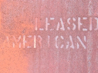 The image features a close-up view of a rusted metal surface with partially legible words stenciled in white, appearing to spell 'LEASED' and 'AMERICAN.' The texture of the metal is rough, with a weathered and corroded appearance, giving it an industrial and aged look.