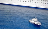 A small pilot boat navigates alongside a large white cruise ship. The ocean water is a rich blue, contrasting with the bright hull of the ship. The pilot boat is labeled 'PILOTO' and is equipped with several antennas and safety equipment.