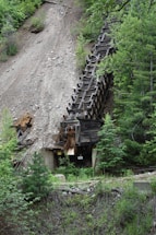 An abandoned, old wooden mining structure is situated on a steep, rocky hillside, partially surrounded by lush green foliage and trees.